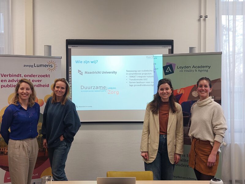 Workshop realistische evaluatie, vertegenwoordigers van awpg lumens en de onderzoekers van Universiteit Maastricht die de workshop gaven voor hun banners met het scherm dat werd gebruikt voor het geven ervan tussen hen in.